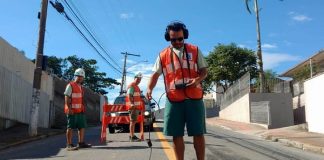 funcionário da casan usando colete laranja usa equipamento de escuta no chão de asfalto molhado; outros dois funcionários atrás na rua;