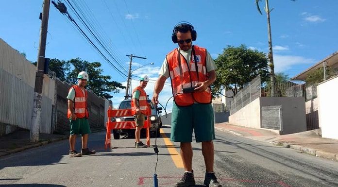 funcionário da casan usando colete laranja usa equipamento de escuta no chão de asfalto molhado; outros dois funcionários atrás na rua;