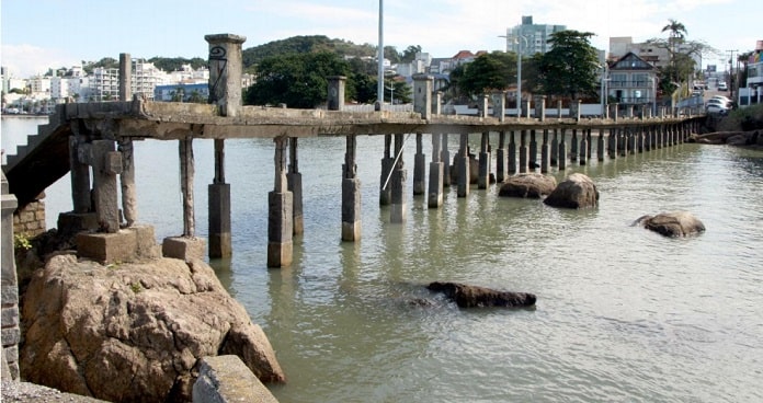 trapiche visto de lado com as palafitas comprometidas; pedras na água; bairro ao fundo