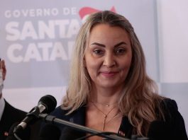 daniela reinehr em frente a um microfone com painel de logos ao fundo