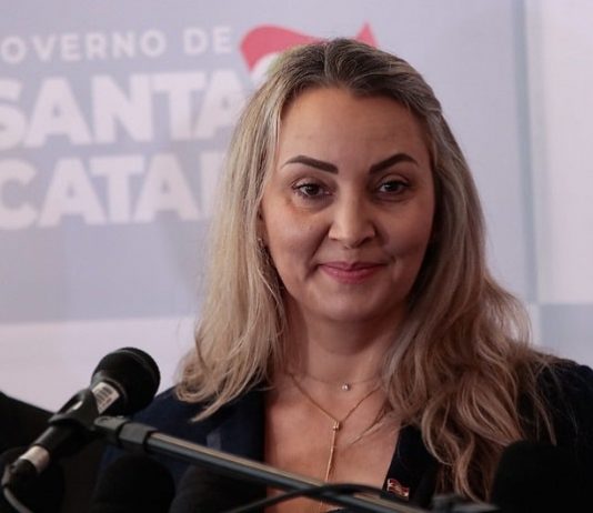 daniela reinehr em frente a um microfone com painel de logos ao fundo