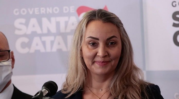 daniela reinehr em frente a um microfone com painel de logos ao fundo