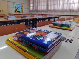 livros e cadernos organizados em pilha sobre carteira em sala de aula com mais mesas e carteiras organizadas; persianas fechadas