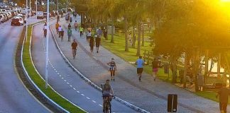 pessoas andando a pé e de bicicleta no calçadão da beira-mar norte, vistas de cima; luz lateral de pôr do sol; carros ao fundo