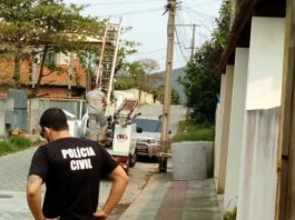 policial usando camiseta com inscrição da polícia civil nas costas em calçada olhando para buraco e ao fundo funcionário da celesc subindo em caminhão ao lado de poste