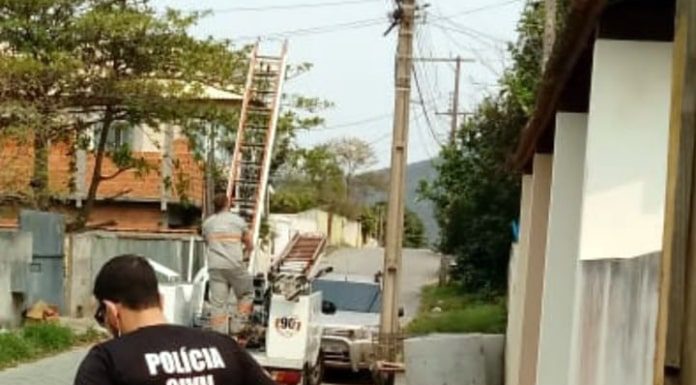 policial usando camiseta com inscrição da polícia civil nas costas em calçada olhando para buraco e ao fundo funcionário da celesc subindo em caminhão ao lado de poste