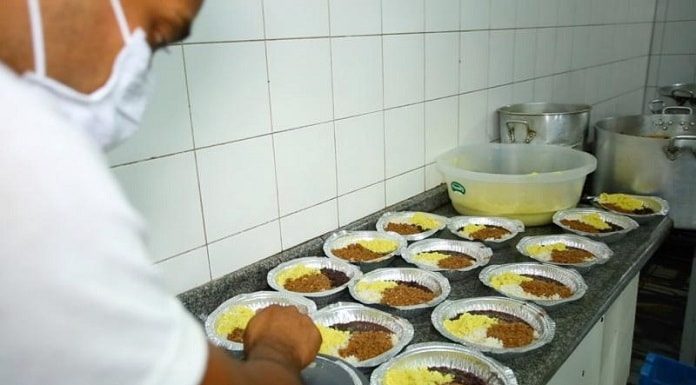 pessoa usando máscara ao lado de balcão com diversos pratos preparados com arroz, feijão e outras comidas em marmitas de alumínio