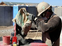 trabalhador da construção civil usando máscara, capacete e óculos de proteção faz preparo de cimento em cima de bancada em área de construção
