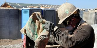 trabalhador da construção civil usando máscara, capacete e óculos de proteção faz preparo de cimento em cima de bancada em área de construção