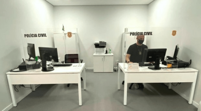duas mesas de escritório com computadores dentro de uma pequena sala; paineis acrílicos transparentes nas mesas com inscrição da polícia civil; um homem sentado atrás da mesa da direita