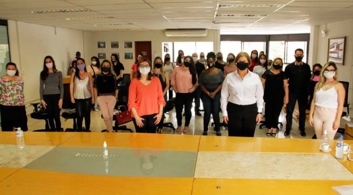 adeliana de dezenas de outras mulheres em pé usando máscaras, distanciadas, posando para foto; grande mesa em primeiro plano