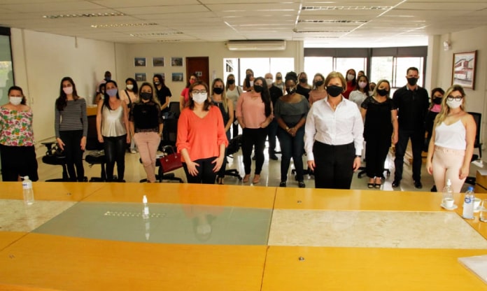 adeliana de dezenas de outras mulheres em pé usando máscaras, distanciadas, posando para foto; grande mesa em primeiro plano