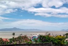 céu com nuvens sobre a baía sul da grande florianópolis; muros, plantas e telhados em primeiro plano; ilha ao fundo