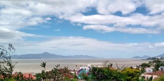 céu com nuvens sobre a baía sul da grande florianópolis; muros, plantas e telhados em primeiro plano; ilha ao fundo