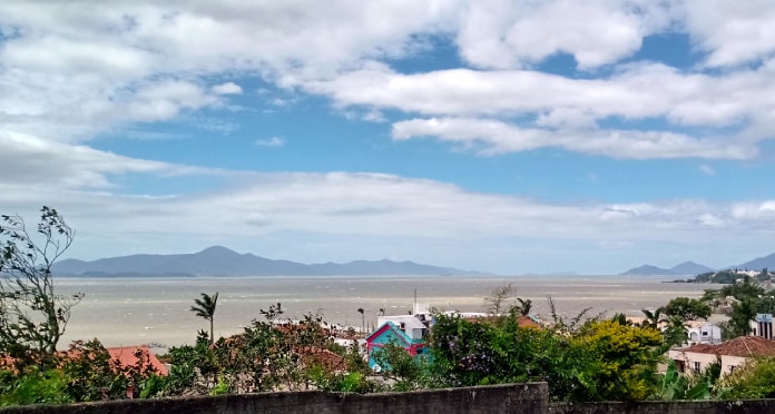 céu com nuvens sobre a baía sul da grande florianópolis; muros, plantas e telhados em primeiro plano; ilha ao fundo