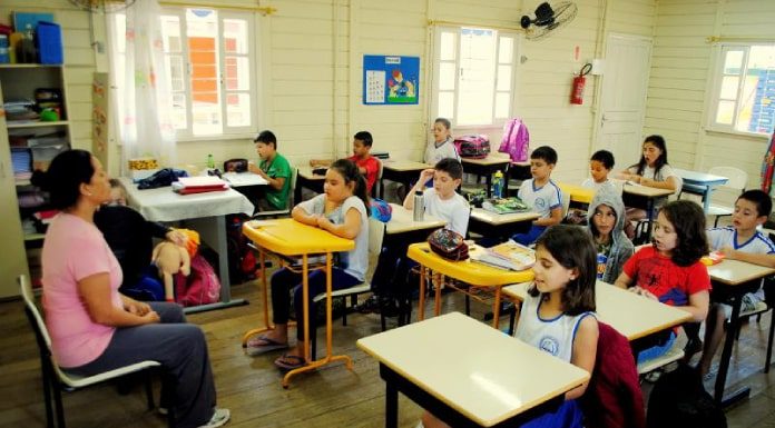 Crianças sentadas em carteiras de sala de aula olhando para professora