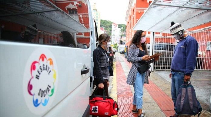 lateral de uma van com inscrição resgate social; na calçada, duas mulheres usando máscaras conversam com homem morador de rua que está de máscara em pé segurando mochila e de roupas de frio