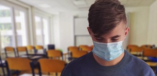 aluno usando máscara lê um livro de frente para a foto; cadeiras de sala de aula ao fundo
