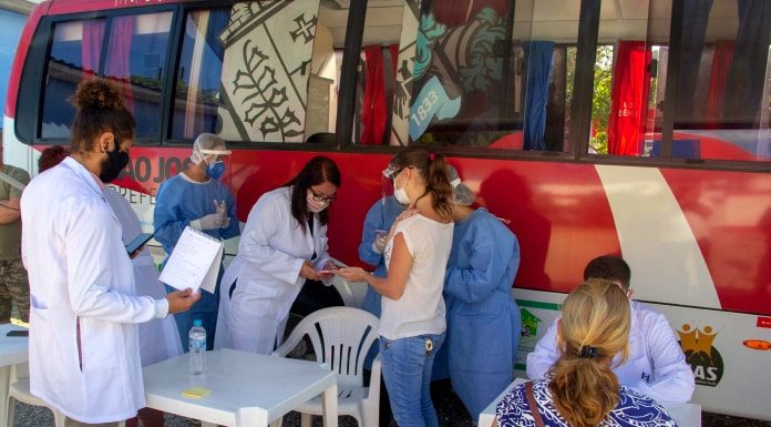 profissionais de saúde e pessoas ao lado de ônibus do município; há cadeiras e mesas de plásticos dispostas no local; maioria das pessoas em pé