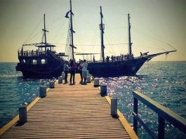 escuna ancorada ao final de um trapiche com quatro pessoas em pé na frente e mais dentro do barco; mar encrespado