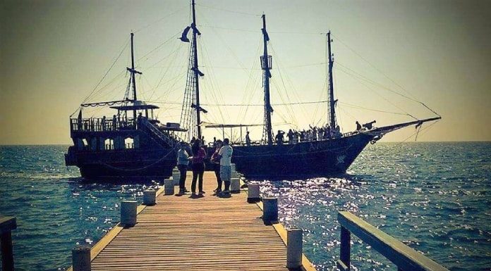 escuna ancorada ao final de um trapiche com quatro pessoas em pé na frente e mais dentro do barco; mar encrespado