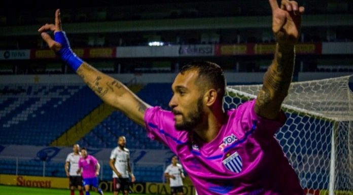 jogador do avaí corre na lateral de campo com os braços levantados comemorando usando camisa rosa; gol e campo com joadores ao fundo; estádio vazio