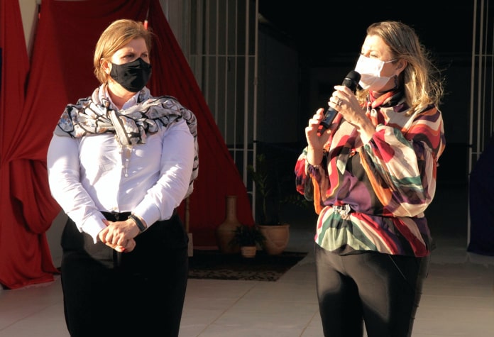 adeliana usando máscara olhada para outra mulher discursando ao microfone usando máscara; ambas em pé
