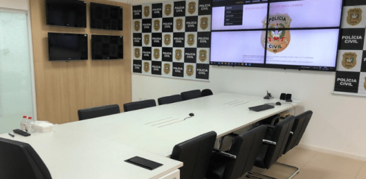 Sala da Polícia Civil com mesa grande, cadeiras e monitores