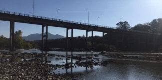 Árvores atrás, na frente uma ponte, logo um rio com pedras que mostra estar em um nível abaixo do devido