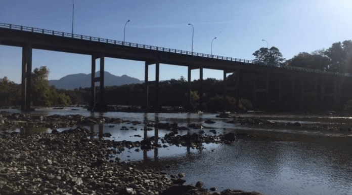 Árvores atrás, na frente uma ponte, logo um rio com pedras que mostra estar em um nível abaixo do devido