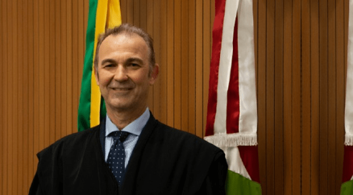 Otávio José Minatto de terno sorrindo, com duas bandeiras atrás