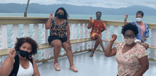 Professoras negras de Florianópolis contam histórias pessoais e desafios de vida Cinco professoras negras de máscara e de punhos cerrado para cima, em sinal de resistência negra