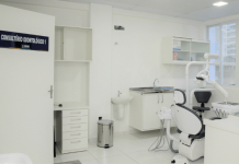 Consultório odontológico da UBS em São José. No lado esquerdo uma porta branca com uma placa azul informando o consultório, mais para a direita atrás aparece uma lixeira, uma piá, armário com equipamentos médicos e na frente uma cadeira para os pacientes.