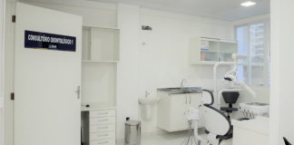 Consultório odontológico da UBS em São José. No lado esquerdo uma porta branca com uma placa azul informando o consultório, mais para a direita atrás aparece uma lixeira, uma piá, armário com equipamentos médicos e na frente uma cadeira para os pacientes.