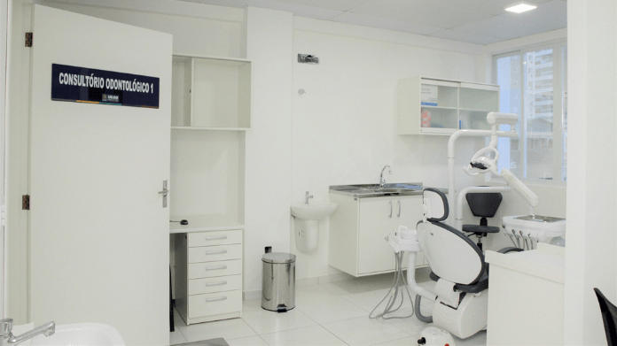 Consultório odontológico da UBS em São José. No lado esquerdo uma porta branca com uma placa azul informando o consultório, mais para a direita atrás aparece uma lixeira, uma piá, armário com equipamentos médicos e na frente uma cadeira para os pacientes.