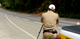 Rodovia catarinense, com policial em pé, de costas para a foto e no canto direito o carro da Polícia Militar