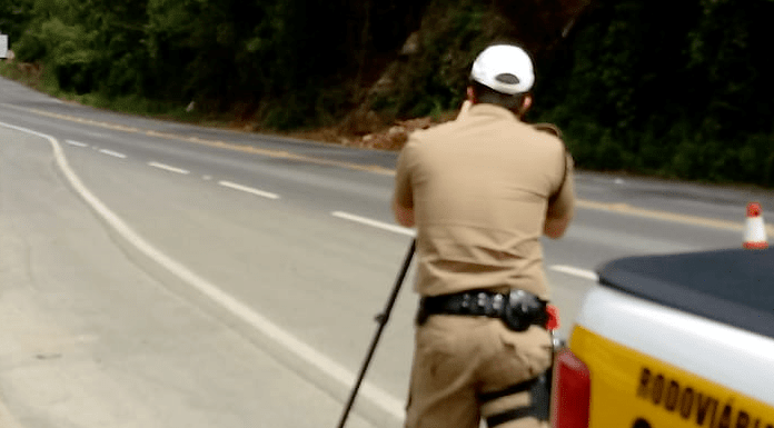 Rodovia catarinense, com policial em pé, de costas para a foto e no canto direito o carro da Polícia Militar