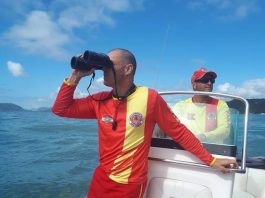 em uma lancha, bombeiro usa binóculos olhando para o mar, outro ao fundo pilota o barco