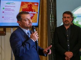 moisés fala ao microfone; atrás uma tela com apresentação e secretário jeferson ao lado