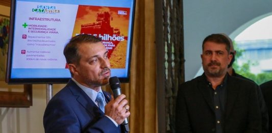 moisés fala ao microfone; atrás uma tela com apresentação e secretário jeferson ao lado