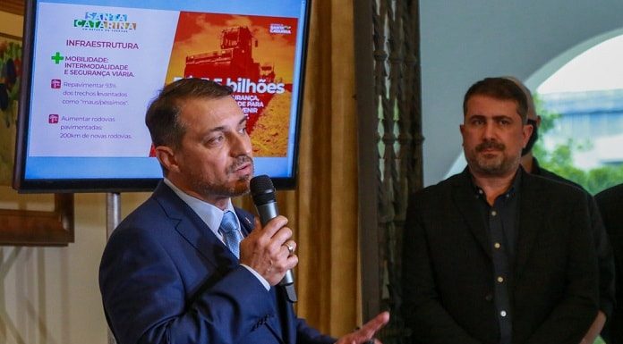 moisés fala ao microfone; atrás uma tela com apresentação e secretário jeferson ao lado