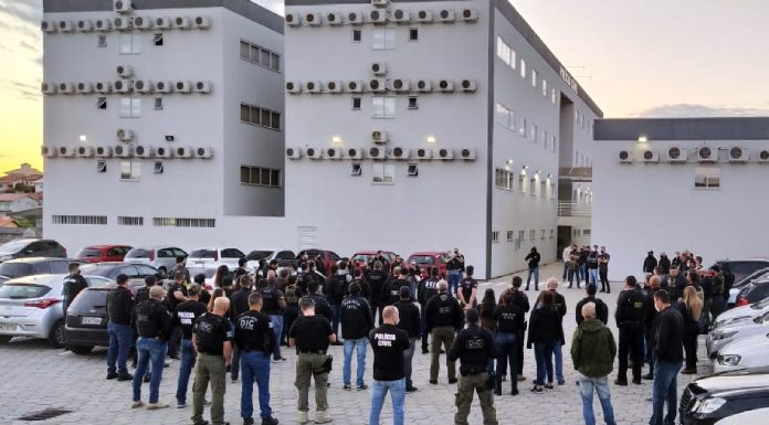 grande grupo de policiais reunidos no estacionamento de delegacia; carros em volta; prédio de 3 andares ao fundo