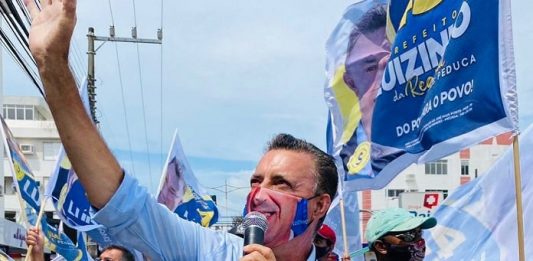 luizinho segura microfone e olha para o alto com a mão para cima; usa uma máscara com seu sorriso; correligionários em volta de máscara e com bandeiras