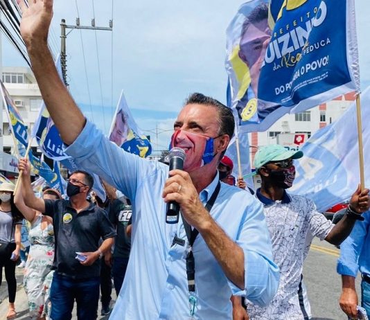 luizinho segura microfone e olha para o alto com a mão para cima; usa uma máscara com seu sorriso; correligionários em volta de máscara e com bandeiras