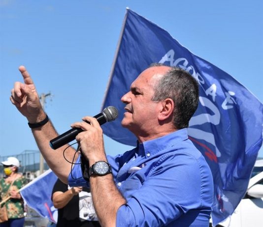 moacir visto de lado discursando ao microfone com dedo em riste; bandeira com seu número ao fundo