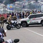 carro jeep da polícia militar atressado na pista com moto lado; motoqueiros em primeiro plano no trânsito. ao fundo no centro da foto grupo protestando e alguns policiais em volta; morro com casas ao fundo