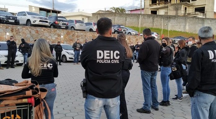 Mecanismo verde: diversos policiais civis homens e mulheres com jaquetas da corporação reunidos em pé em estacionamento; maioria de costas para foto