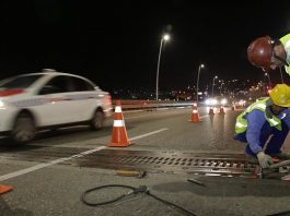 Ponte Pedro Ivo Campos terá fechamento parcial para manutenção dois trabalhadores usando vestimentas de proteção aparafusam placa de metal no chão da ponte com cones em volta, à noite, e taxi passando ao lado