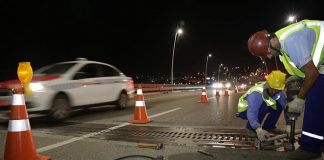 dois trabalhadores usando vestimentas de proteção aparafusam placa de metal no chão da ponte com cones em volta, à noite, e taxi passando ao lado