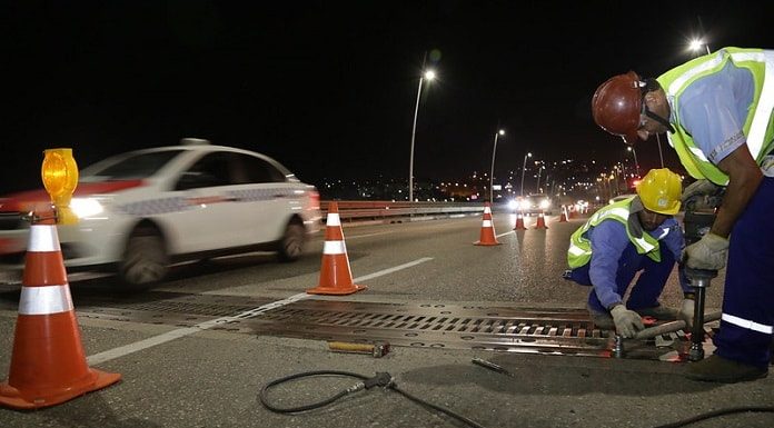 dois trabalhadores usando vestimentas de proteção aparafusam placa de metal no chão da ponte com cones em volta, à noite, e taxi passando ao lado